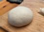 A ball of pizza dough sitting on wood with a kitchen scale in the background.